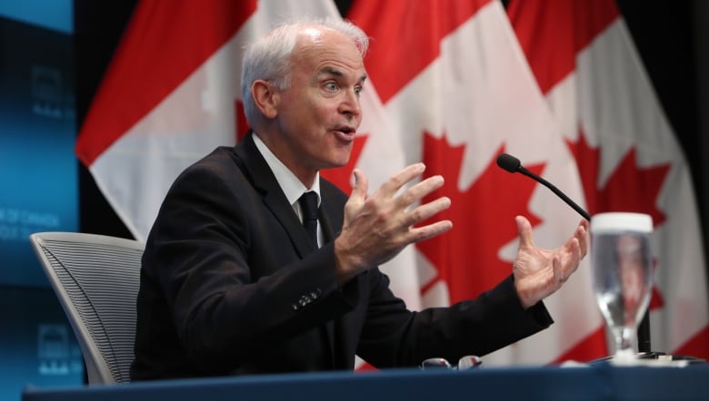 Un hombre vestido con un traje oscuro gesticula ante un micrófono, frente a banderas canadienses.