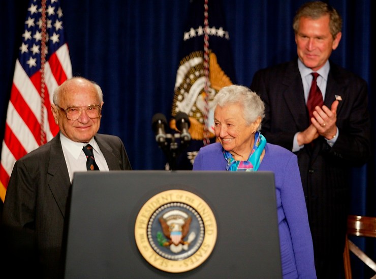 Milton y Rose Friedman están detrás de una tribuna con el sello presidencial.  El presidente de los Estados Unidos, George Bush, aplaude al fondo.