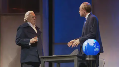 BBC Brian Cox en el escenario presenta a Adam Smith. Lleva una peluca rubia/blanca, una chaqueta oscura y una camisa salvaje. Habla con un hombre con traje y gafas y hay una mesa y un globo en el escenario  