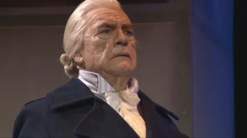 Brian Cox en el escenario presenta a Adam Smith. Lleva una peluca rubia/blanca, una chaqueta oscura y una camisa salvaje.