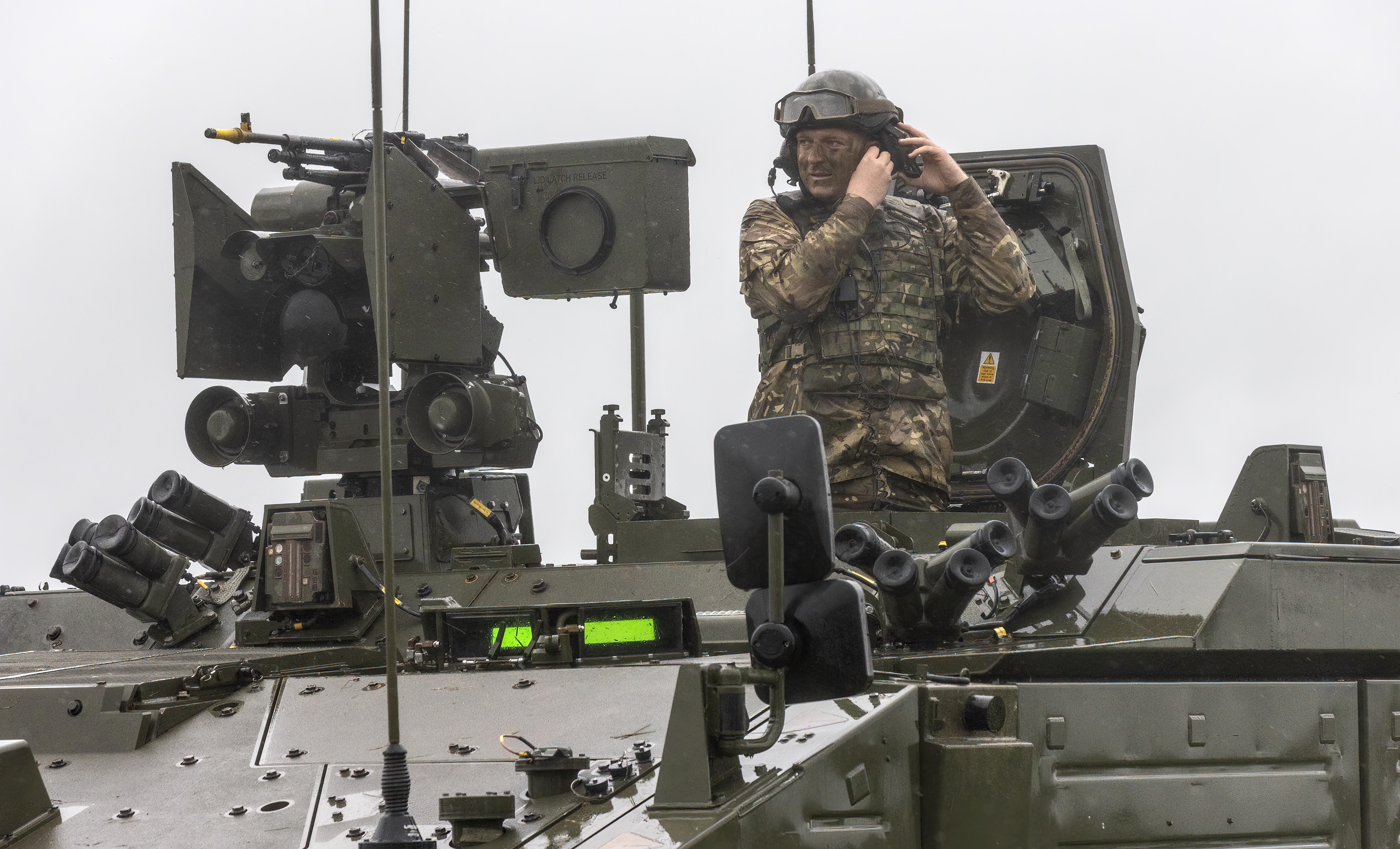 Un soldado británico con camuflaje y casco, cubierto de barro, mira por la puerta de un vehículo de combate blindado de la serie Ajax.