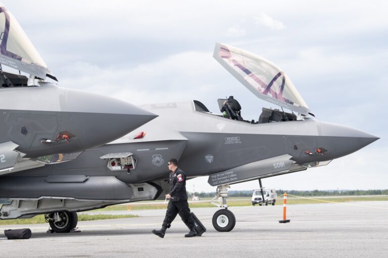 En esta toma se pueden ver el morro cónico y las escotillas abiertas de dos aviones de combate F-35. Los aviones están estacionados en la pista y un tripulante de tierra con un mono y gafas de sol pasa arrastrando un juego de herramientas con ruedas.