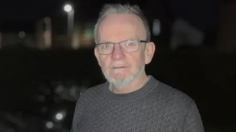 BBC Ciarán O'Doherty lleva un jersey estilo armario. Está parado en un punto de carga de vehículos eléctricos, es de noche y muy oscuro.