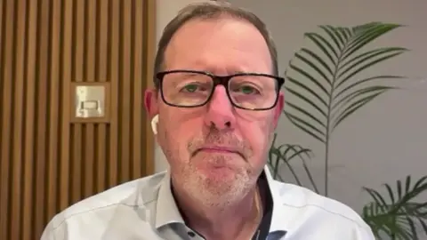 Mark McCall tiene cabello corto color castaño claro, usa anteojos con montura negra y una camisa de color claro.
