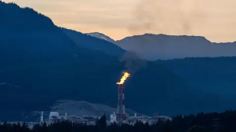 Bloomberg vía Getty Images Una pila de bengalas blancas y rojas envía fuego al cielo nocturno en las instalaciones de LNG Canada en Kitimat, Columbia Británica, Canadá. Al fondo hay montañas adyacentes a un cielo de color naranja pálido. 