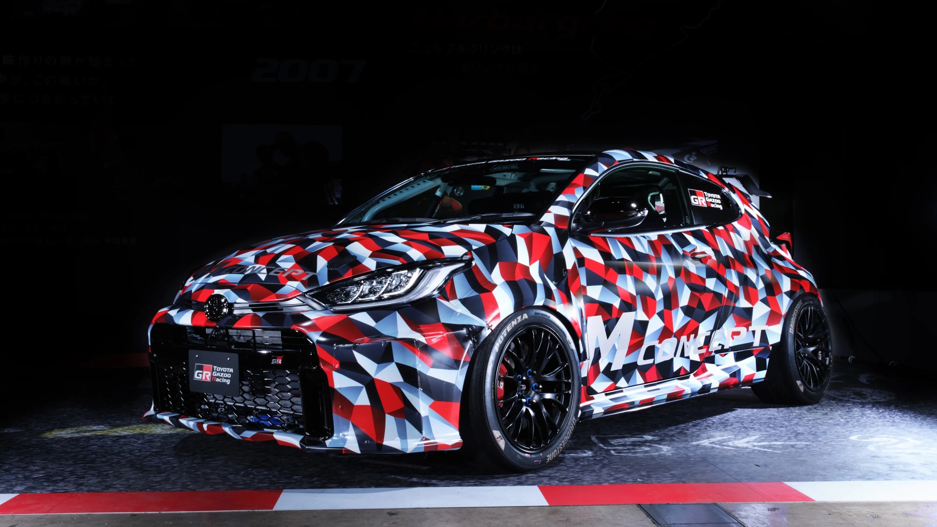Coche de carreras Toyota GR con camuflaje geométrico rojo, blanco y negro y ruedas negras sobre un suelo oscuro.